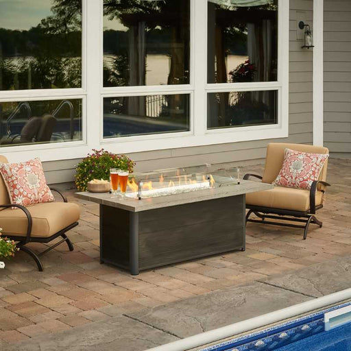 The Outdoor Greatroom Cedar Ridge Linear Gas Fire Pit Table near Pool with Clear Tempered Fire Glass Gems, Fire Burner On and Glass Wind Guard